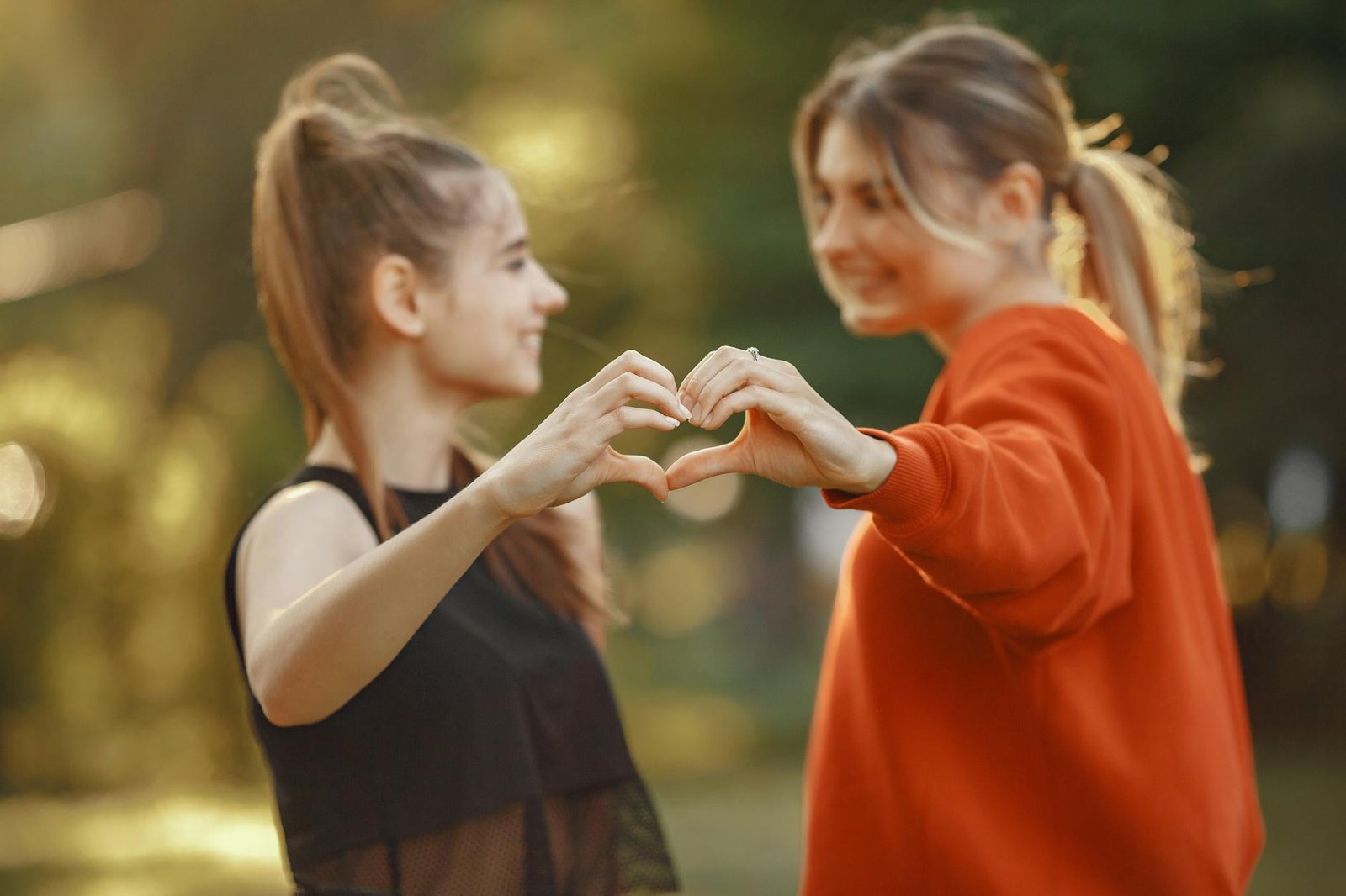 Two young women forming a heart shape with hands in a sunny park setting, symbolizing friendship and love.