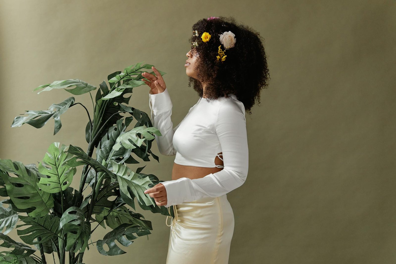 Stylish woman with afro hair and floral decor poses among lush green plants, showcasing modern fashion.