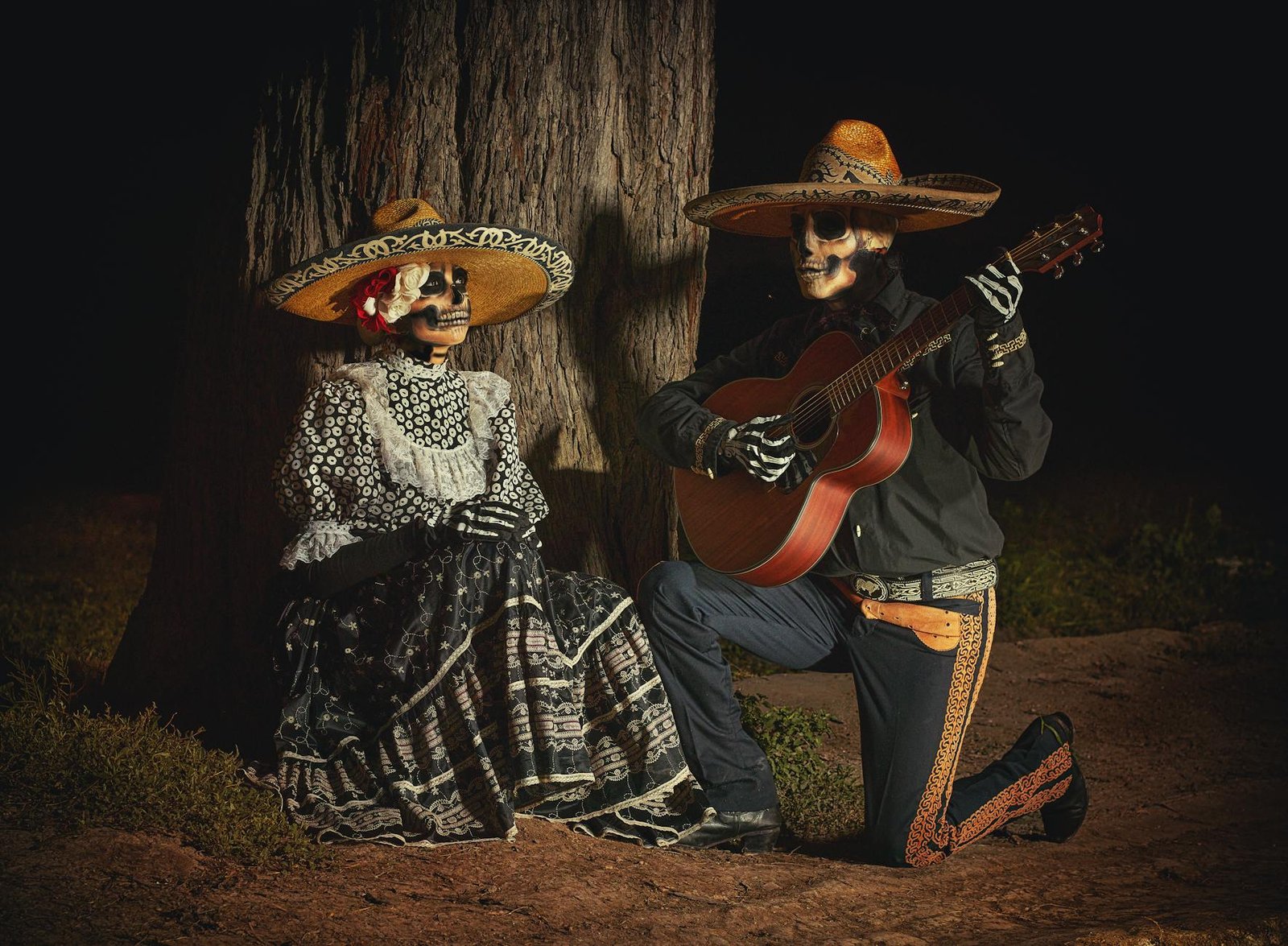 Traditional Day of the Dead themed musicians with skull makeup in Mexican attire by a tree.