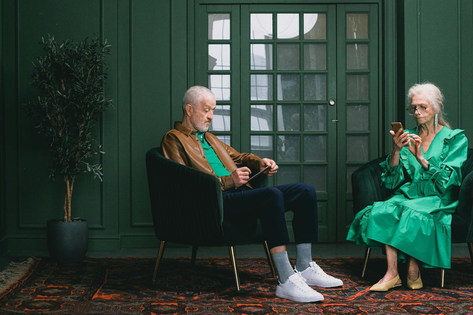 Senior man and woman using smartphones in a stylish green room with armchairs and plant.