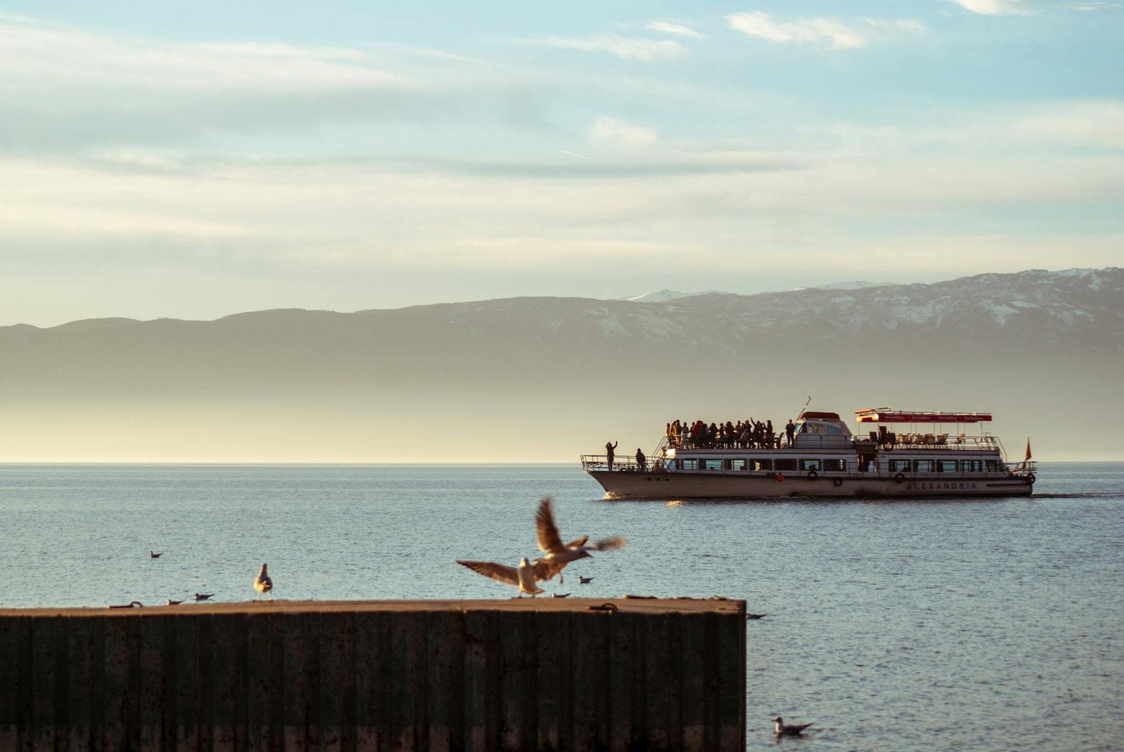 A scenic view of a boat cruising on Lake Ohrid at sunset with birds flying around.